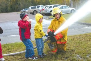 Staff photos/Rhonda Silence The Grand Marais Fire Department visited Sawtooth Elementary and Great Expectations in Grand Marais on Wednesday, October 7 during Fire Prevention Week. Firefighters Ben Silence and Gideon Silence from the Grand Marais Fire Department and Kim Linnell, Clark Bloomquist, and Paul McFarlane from Colvill Fire Department talked about fire safety and demonstrated hoses and fire equipment. Above: Tanner Berglund excitedly waits for his turn as Riley Goettl shoots the fire hose with firefighter Gideon Silence. Right: Fireman Paul McFarlane of the Colvill Fire Department explains the various hoses on the bright green Colvill fire truck.