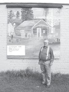 Staff photo/Jane Howard Lyle Saethre in front of his mural of the Omar Ellquist log cabin on Maple Hill. Saethre has been painting murals of historic North Shore scenes all over Grand Marais.