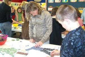 Students at Birch Grove Community School have been hard at work, researching, designing and arranging tiles for a mosaic to be placed on an exterior wall of the Birch Grove Community Center in Tofte. Above left: Madysen McKeever, 11, and Jared DeHart, 11, work on laying the tiles for a dog musher and pine tree. Lower left: A mural at Birch Grove of course needs birch trees! Jacob Bjerkness, 10, carefully places the white and black tiles to create a birch tree. Lower right: Kindergartener Nathan Bilben adds blue tiles to give a moose an eye.