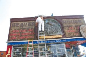 Touch-up time! Above: Yarrow Korf checks out the sculpture on the front of the Beaver House in downtown Grand Marais. Korf and Jim Baird specialize in sandblasting and restoration of buildings, but this project has special meaning—Yarrow’s grandfather, Jim Korf, created the beaver dam. Far left: The giant walleye, also created by Jim Korf, was kept under cover as work was being done. Left: Jim Baird works on the soon-tobe chartreuse lettering for the front of the building.