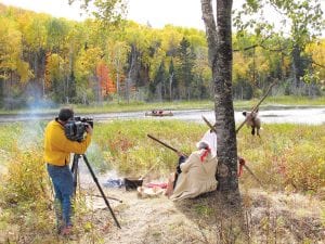 Behind the camera is Chris Wheeler, producer for Great Divide Pictures, shooting a scene for Rendezvous with History: A Grand Portage Story. The scene features Ernest Boyes of Grand Portage on shore as Mike Cyrette and Sarah Deschampe, also of Grand Portage, paddle the canoe.