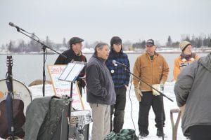 Above: The “Sled Dogs to St. Paul” rally in Grand Marais Harbor Park included words from John Morrin of Grand Portage, vicechairman of the Grand Portage Band of Lake Superior Chippewa. Inset right: Marco Good and Yvonne Mills sang two songs. They encouraged the crowd to sing along. Upper right: Especially meaningful was the participation of the Stonebridge Singers of Grand Portage. Lower right: A kiss for luck. Sherrie Moe gives her husband Frank Moe a kiss just before he takes off on his almost 400-mile journey to St. Paul.