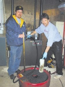 Cook County Schools Maintenance Mechanic Tom Nelson (left) and Facilities and Transportation Director Mike Groth in front of the school bus garage furnace that is fueled by used motor oil. They are pumping fuel into the furnace from an air-powered oil handler that collects oil from school buses. Other sources of oil are the Cook County Highway Department, the City of Grand Marais’ power plant, and Steve’s Sports. The buses themselves provide about 28 days of heat for the building each year.