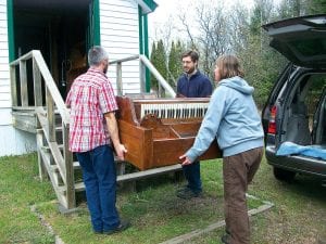 Above: Thanks to the generous volunteers and donors, the organ that once played hymns at church services at the Chippewa City Church in Grand Marais has been returned. Carefully bringing the old organ back to the Chippewa City Church are (LR) Michael McHugh, Museum Director Carrie McHugh and Sam West.