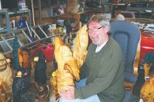 Chainsaw artist Wayne Johnson sits with an eagle he is working on, some of his recent work surrounding him. Only two years into his craft, Wayne is already quite accomplished. His mother Jean asked Wayne to make a carving of her dog. She also asked him to carve a curling stone.